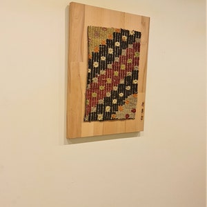 Puede incluir: Una pieza de tela tejida enmarcada con un patr&oacute;n geom&eacute;trico en tonos de rojo, negro, blanco y beige. La tela est&aacute; montada en un marco de madera claro.