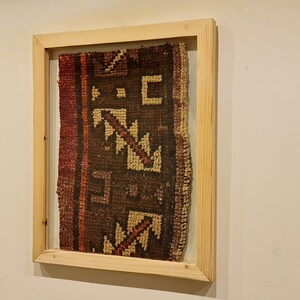May include: A framed piece of fabric with a geometric pattern in shades of brown, red, and beige. The fabric is woven with a traditional design and is displayed in a simple wooden frame.