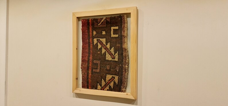 May include: A framed piece of fabric with a geometric pattern in shades of brown, red, and beige. The fabric is woven with a traditional design and is displayed in a simple wooden frame.