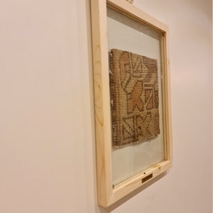May include: A framed piece of woven fabric with a geometric pattern in shades of brown, beige, and white. The frame is made of natural wood and has a glass front.