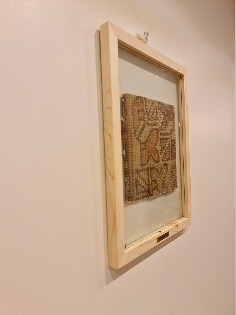 May include: A framed piece of woven fabric with a geometric pattern in shades of brown, beige, and white. The frame is made of natural wood and has a glass front.