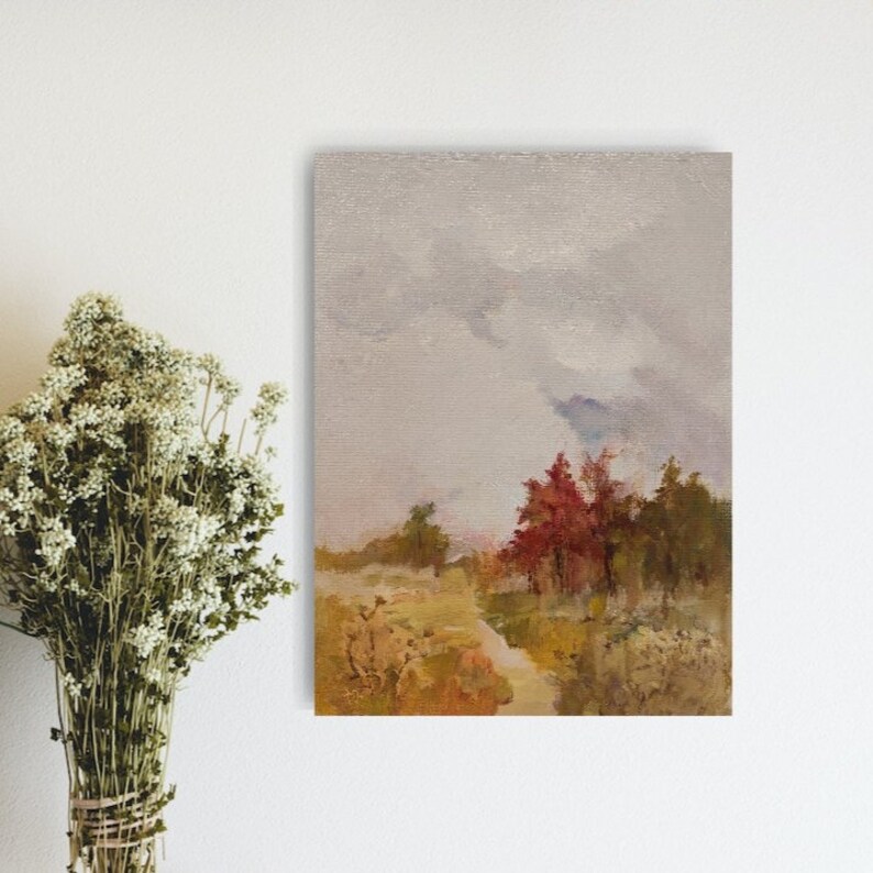 May include: An oil painting depicts a landscape with a path leading towards a cluster of trees with red and green foliage under a cloudy sky. The artwork is framed and mounted on a white wall, with a bundle of dried flowers to the left.