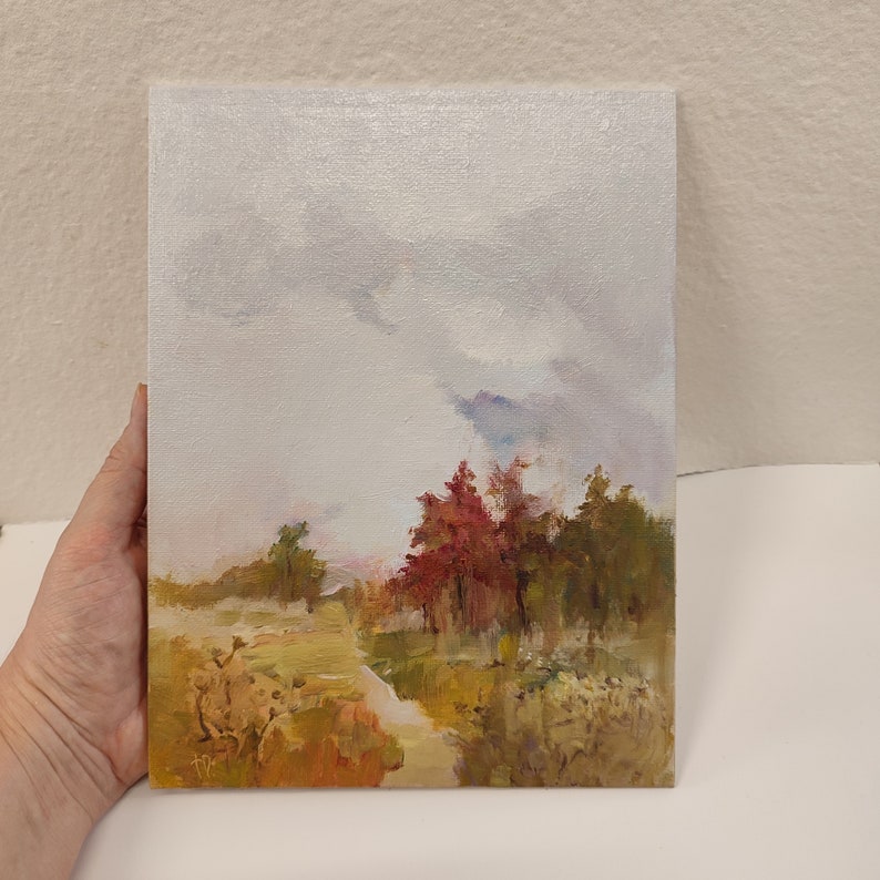 May include: An original oil painting on canvas depicting a landscape scene. The artwork features a cloudy sky above a forest with trees in shades of red, green, and brown. The painting is held by a hand.