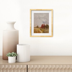 May include: A framed landscape art print with a gold-colored frame. The artwork depicts a field with trees in shades of red and green under a cloudy sky. The print is displayed on a beige cabinet with decorative vases and a small succulent plant.