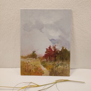May include: Oil painting on canvas depicting a landscape scene. The artwork features a path leading to a cluster of trees with red and green foliage under a cloudy sky. The painting has a soft, impressionistic style with muted colors.