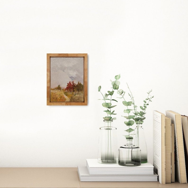 May include: A framed landscape painting with a wooden frame. The artwork depicts a path leading through a field with trees in shades of red and green under a cloudy sky. The painting is displayed on a white wall with decorative vases and books.