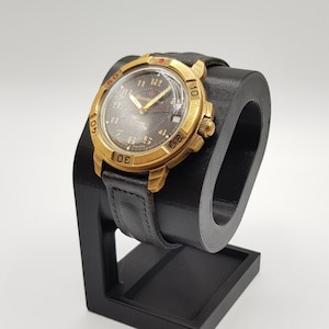 Può includere: Un orologio da polso dorato con quadrante nero e cinturino in pelle nera. L'orologio è su un supporto per orologi nero.