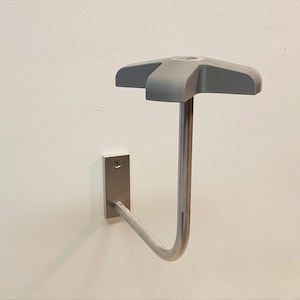 Puede incluir: Un soporte para casco de pared plateado con un reposacascos de plástico gris. El soporte tiene forma de J y un extremo redondeado. El reposacascos tiene forma de estrella de cinco puntas.