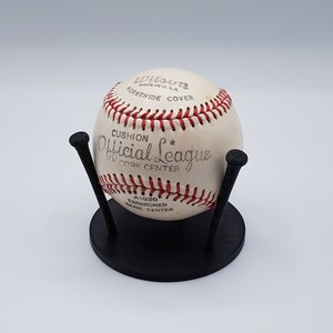 Puede incluir: Una pelota de béisbol vintage con costuras rojas y el texto "Official League" y "Wilson Made in U.S.A." se muestra en un soporte negro. La pelota de béisbol tiene una cubierta de cuero blanco y está diseñada para el deporte del béisbol.