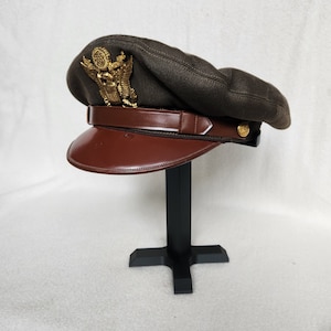 Puede incluir: Un sombrero marrón de estilo militar con un emblema de águila dorada y una visera de cuero marrón. El sombrero se muestra en un soporte negro.