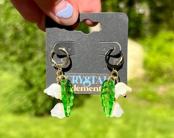 May include: Gold hoop earrings with green leaf and white flower charms. The earrings are hanging from a black tag that reads "CRYSTAL element". The background is a blurred green.