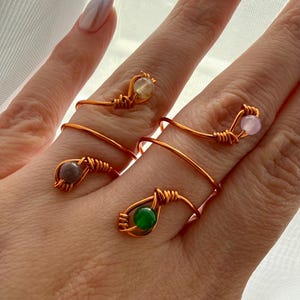 May include: Four copper wire rings with different colored gemstone accents. The gemstones are yellow, pink, gray, and green. The rings are wrapped around the fingers of a hand. The rings are handmade and have a unique, artistic design.