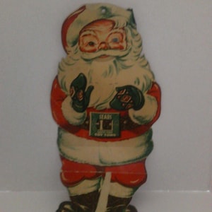 Könnte beinhalten: Vintage Weihnachtsschmuck mit Weihnachtsmann. Die Santa-Figur trägt einen roten Anzug mit weißem Besatz, schwarzen Stiefeln und grünen Handschuhen. Der Text "SEARS TOY TOWN" ist auf dem Gürtel zu sehen. Die Dekoration besteht aus Pappe und hat oben ein Loch zum Aufhängen.