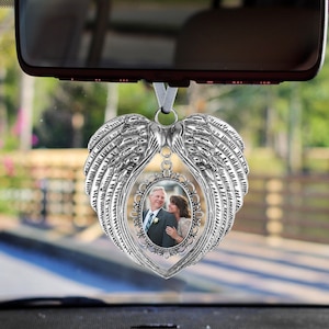May include: Silver car charm featuring a photo frame shaped like angel wings. The frame holds a photo of a couple.
