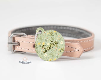 May include: A round, green and white floral dog tag with the name "Josie" written in black cursive. The tag is attached to a brown leather dog collar.