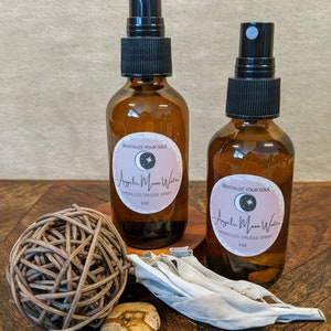 May include: Two amber glass bottles with black spray tops and labels that read "Angelic Moon Water" and "Smokeless Smudge Spray" on a wooden surface with a white sage bundle and a small brown stone.
