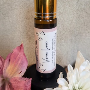 Puede incluir: Un pequeño frasco de vidrio con una tapa dorada y una etiqueta rosa que dice "Venus Love" en una fuente decorativa. La etiqueta también incluye el texto "Essential Oil Roll-on Blend", "Love Healing", "Aphrodisiac", "Reiki Infused" y "Spiritual Well-being".