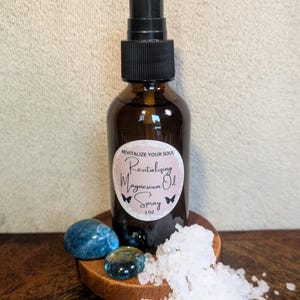 Puede incluir: Una botella de vidrio marrón con un pulverizador negro y una etiqueta que dice "Revitalize Your Soul Revitalizing Magnesium Oil Spray 2 oz". La botella está sobre una superficie de madera con sal blanca y piedras azules.