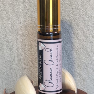 May include: A brown glass roll-on bottle with a gold cap. The label is pink and black and says "Glamour Guard Essential Oil Roll-on Blend" and "Spiritual & Psychic Attack Protection Ward Off Energy Vampires".
