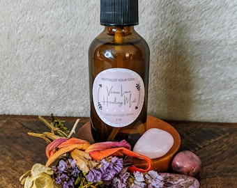 May include: A brown glass bottle with a black spray top and a label that reads "Venus Love Healing Mist" and "Reiki Infused Spray 2 oz". The bottle is sitting on a wooden surface with a pink rose quartz crystal, a pink and white crystal, and a bouquet of dried flowers.