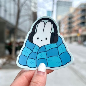 May include: A white bunny wearing headphones is tucked under a blue quilted blanket. The sticker is perfect for adding a touch of whimsy to laptops, water bottles, or journals.