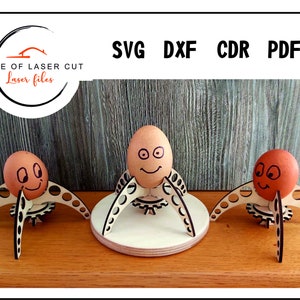 Puede incluir: Tres soportes para huevos de madera con un dise&ntilde;o de engranaje y un huevo marr&oacute;n con una cara dibujada en cada soporte. Los soportes est&aacute;n sobre una superficie de madera.
