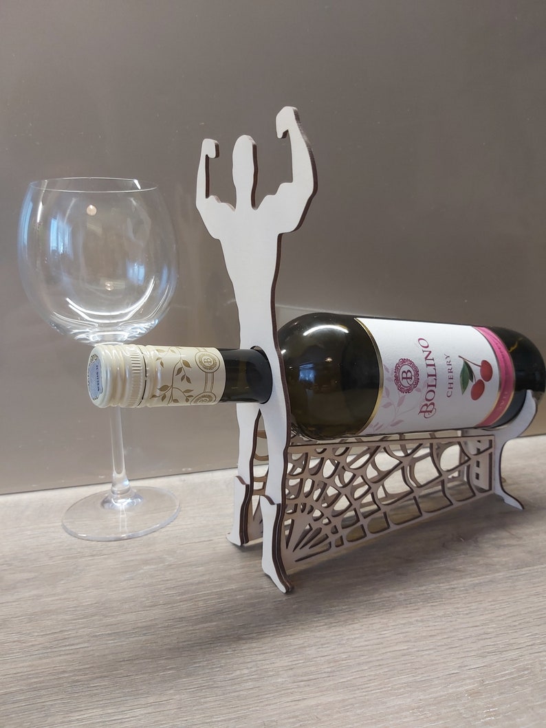 May include: Wooden wine bottle holder shaped like a person with arms raised in a muscle pose. The holder has a spiderweb design and is holding a bottle of Bollino Cherry wine.