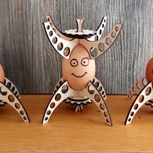 Puede incluir: Tres soportes para huevos de madera con un dise&ntilde;o de engranaje. Cada soporte tiene un huevo marr&oacute;n con una cara sonriente dibujada.