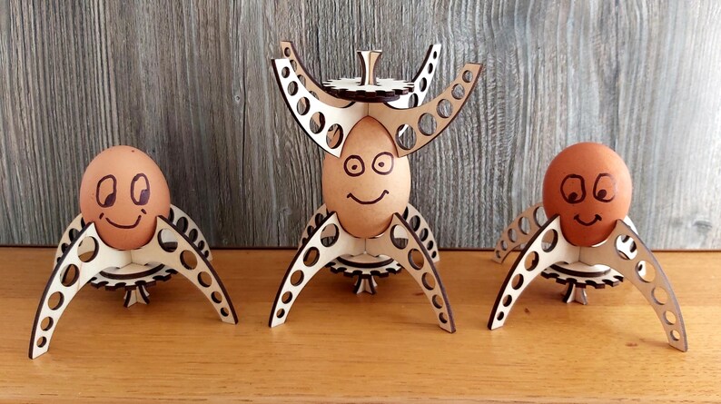 Puede incluir: Tres soportes para huevos de madera con un dise&ntilde;o de engranaje. Cada soporte tiene un huevo marr&oacute;n con una cara sonriente dibujada.