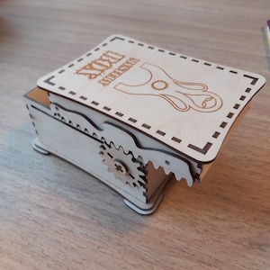 May include: A wooden box with a hinged lid. The lid has a laser-cut design of a slingshot with the text "YOU ARE AMAZING" above it. The box has a gear mechanism on the side.