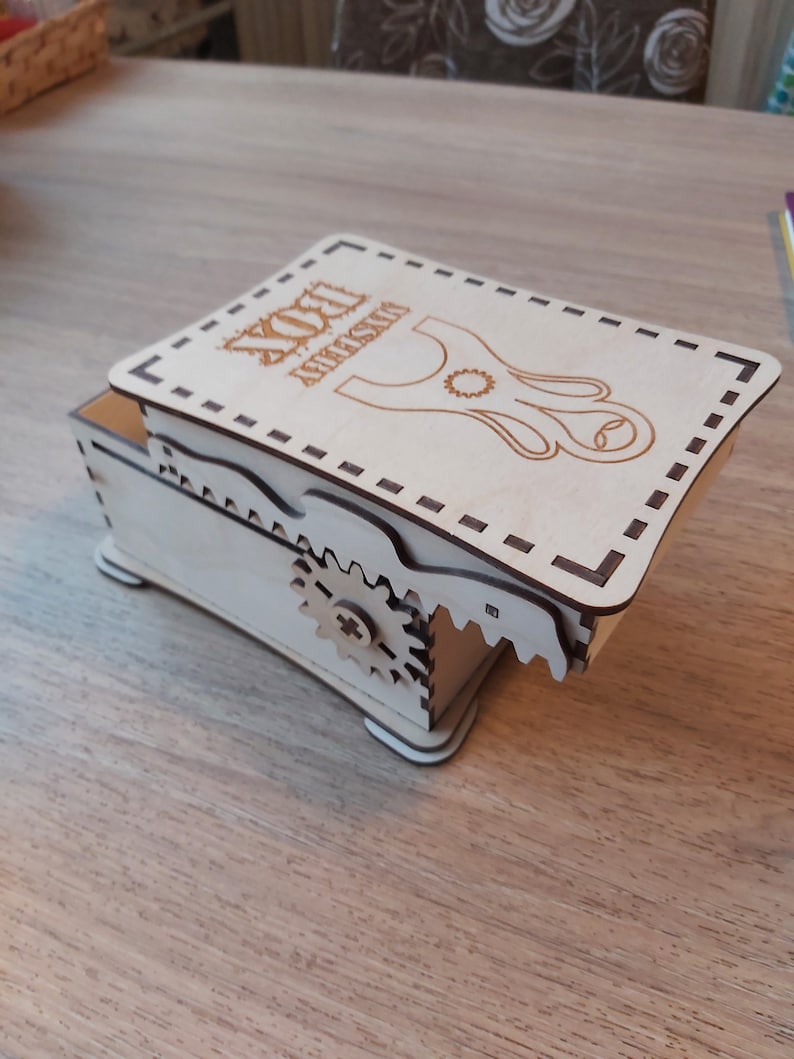 May include: A wooden box with a hinged lid. The lid has a laser-cut design of a slingshot with the text "YOU ARE AMAZING" above it. The box has a gear mechanism on the side.