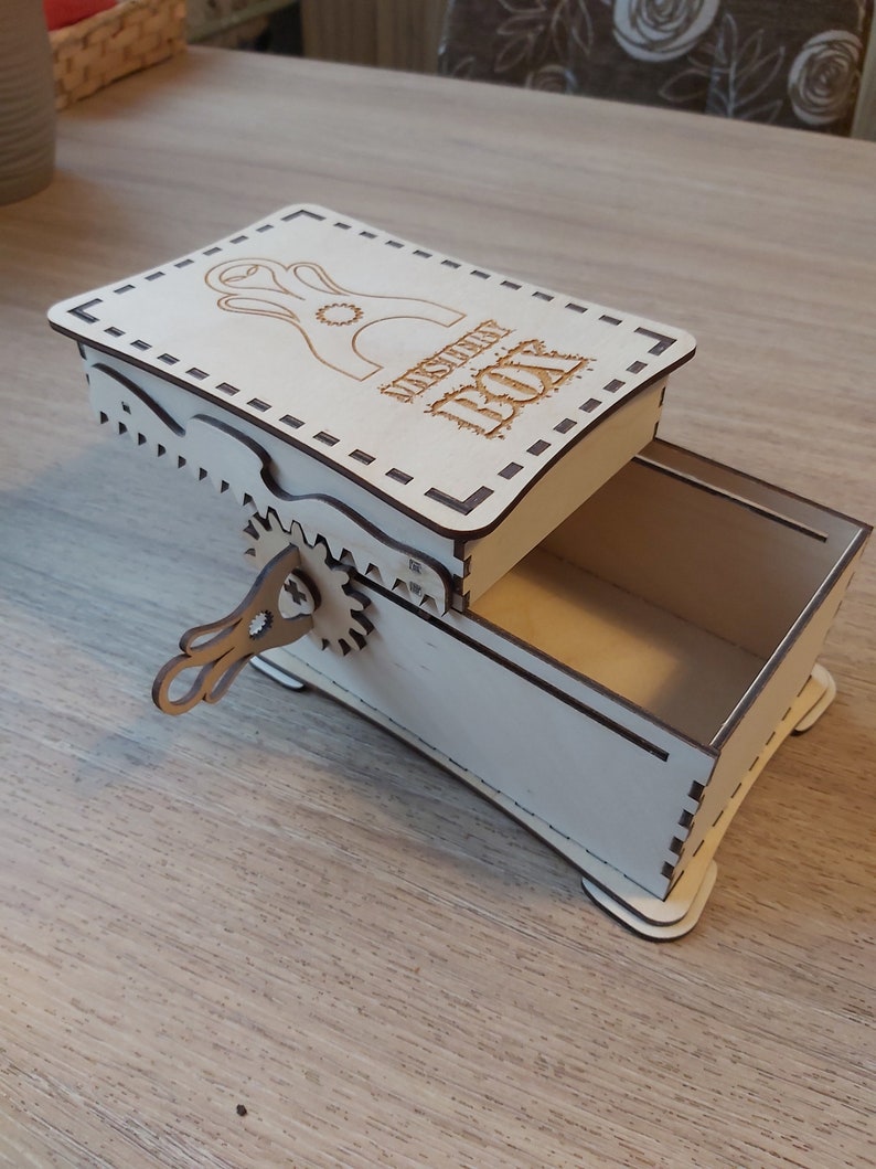 May include: A wooden box with a hinged lid and a small, intricate metal crank on the side. The lid is decorated with a laser-cut design and the words "Mystery Box".