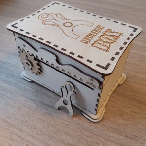 May include: A wooden box with a hinged lid and a small gear on the side. The box is decorated with a laser-cut design and the words "Mystery Box".