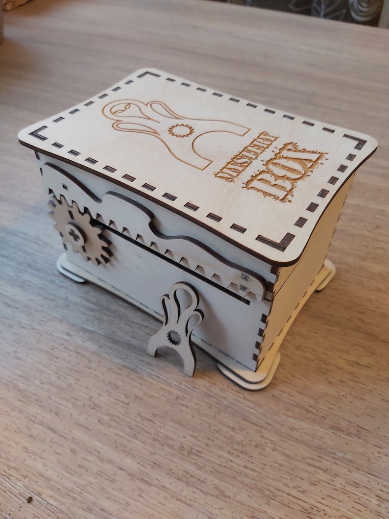 May include: A wooden box with a hinged lid and a small gear on the side. The box is decorated with a laser-cut design and the words "Mystery Box".