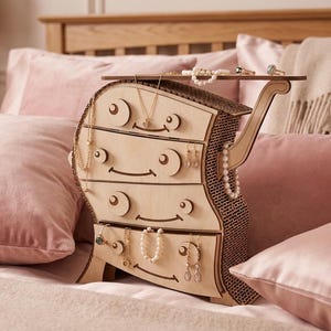 May include: A whimsical wooden jewelry organizer with a unique, curved design. The organizer has four drawers with circular accents and a top shelf, holding necklaces, earrings, and rings. The light-colored wood contrasts with the pink pillows and bedding in the background.