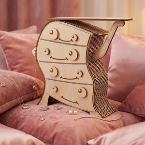 May include: A whimsical wooden jewelry chest with four drawers and a curved design. The chest is light brown and sits on a pink velvet surface, with gold necklaces and rings displayed nearby. The chest has a shelf on top.