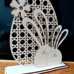 K&ouml;nnte beinhalten: Osterei-Dekoration aus Holz mit zwei Hasen mit langen Ohren und einem gro&szlig;en Ei mit gewebtem Muster. Das Ei ist mit einer Blume verziert.