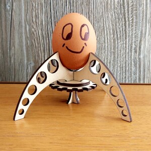 Puede incluir: Un soporte para huevos de madera con una cara sonriente dibujada en el huevo. El soporte est&aacute; hecho de madera cortada con l&aacute;ser y tiene una base circular con un dise&ntilde;o de engranaje.