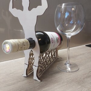 May include: White wooden wine bottle holder shaped like a muscular figure with arms raised. The holder has a spiderweb design on the base and a bottle of wine is resting on the figure's arms.