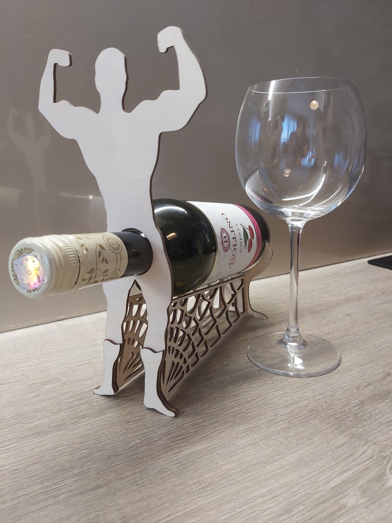 May include: White wooden wine bottle holder shaped like a muscular figure with arms raised. The holder has a spiderweb design on the base and a bottle of wine is resting on the figure's arms.