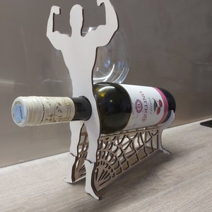 May include: A wooden wine bottle holder shaped like a muscular figure with arms raised. The holder features a cutout design resembling a spider web. A bottle of white wine is resting in the holder.