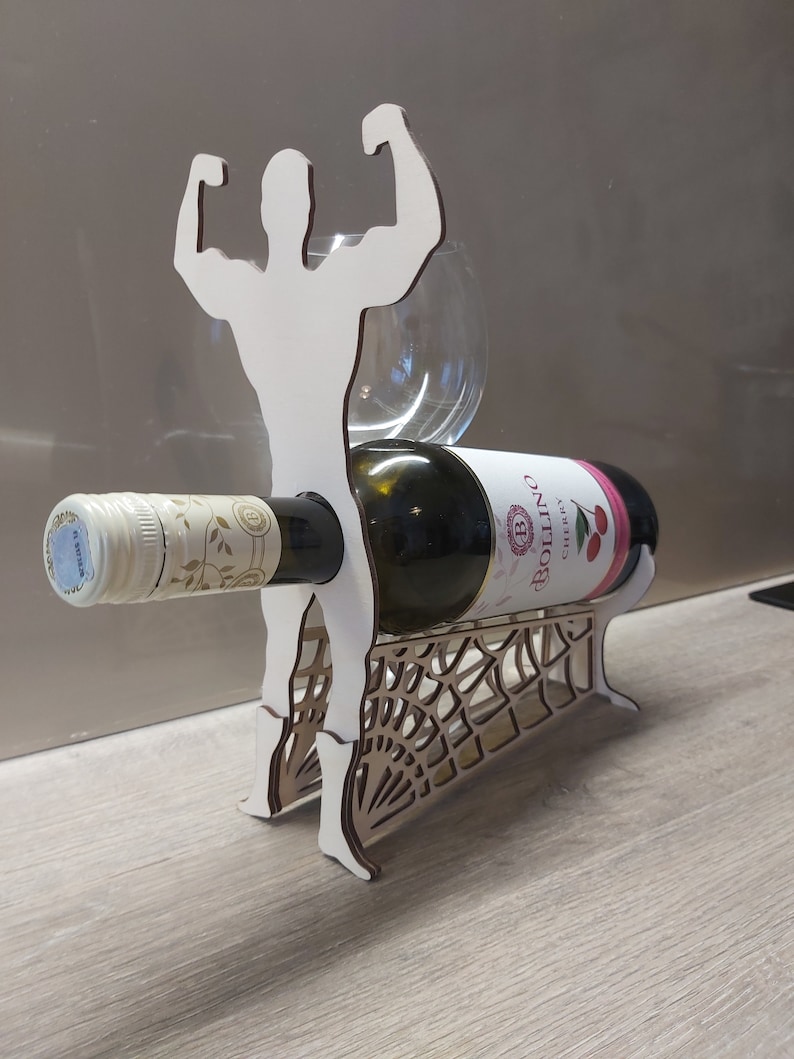 May include: A wooden wine bottle holder shaped like a muscular figure with arms raised. The holder features a cutout design resembling a spider web. A bottle of white wine is resting in the holder.