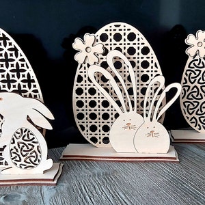 K&ouml;nnte beinhalten: Drei h&ouml;lzerne Ostereier-Dekorationen mit verschiedenen Designs. Das erste Ei hat ein geometrisches Muster und ein Kaninchen. Das zweite Ei hat ein gewebtes Muster und zwei Kaninchen. Das dritte Ei hat ein keltisches Knotenmuster und ein K&uuml;ken.