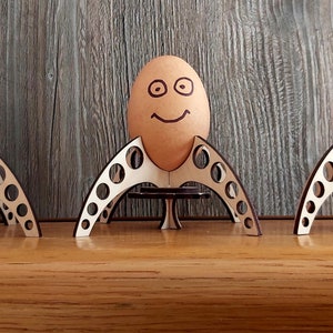 Puede incluir: Tres soportes para huevos de madera con forma de cohetes, cada uno con un huevo marr&oacute;n con una cara sonriente dibujada.