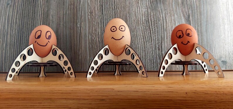 Puede incluir: Tres soportes para huevos de madera con forma de cohetes, cada uno con un huevo marr&oacute;n con una cara sonriente dibujada.