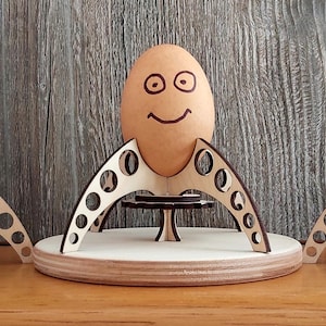 Puede incluir: Tres soportes para huevos de madera con forma de cohetes, cada uno con un huevo marr&oacute;n con una cara sonriente dibujada.