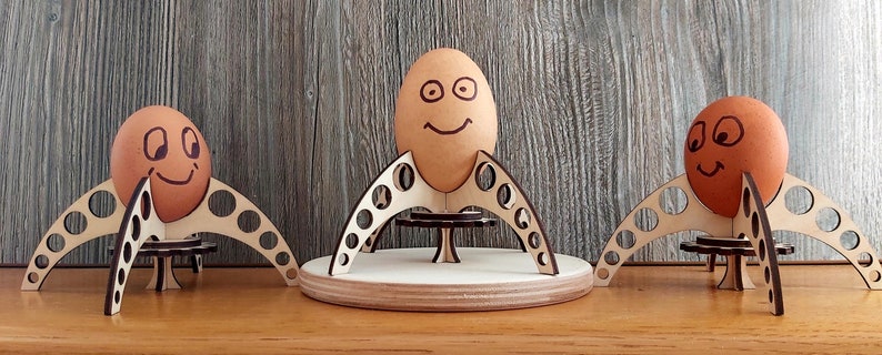 Puede incluir: Tres soportes para huevos de madera con forma de cohetes, cada uno con un huevo marr&oacute;n con una cara sonriente dibujada.