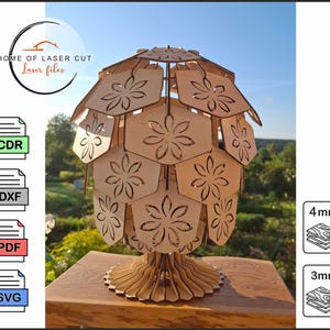 May include: A wooden lamp with a floral design, laser cut from light-colored wood. The lamp has a round base and a shade composed of hexagonal panels with flower cutouts. The image includes the text "HOME OF LASER CUT Laser files".