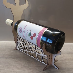 May include: A wooden wine bottle holder shaped like a muscular figure with arms raised. The holder is designed to hold a bottle of wine in a web-like design. The bottle is a dark red wine with a label that reads "Bollino Cherry Sweet & Fruity".