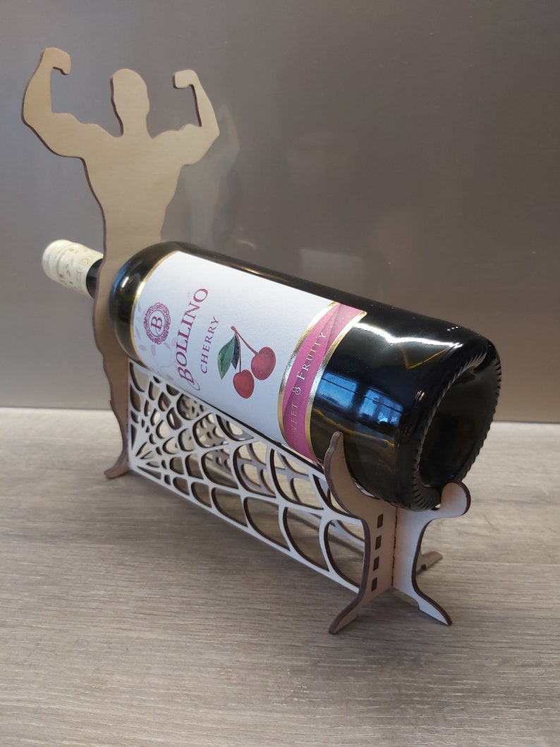 May include: A wooden wine bottle holder shaped like a muscular figure with arms raised. The holder is designed to hold a bottle of wine in a web-like design. The bottle is a dark red wine with a label that reads "Bollino Cherry Sweet & Fruity".